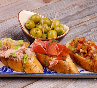 Tapas Bruschetta with Bacon, Vegetables, Cured Ham, Pate on Garlic Bread and Green Olives Snack on Rustic Wooden background