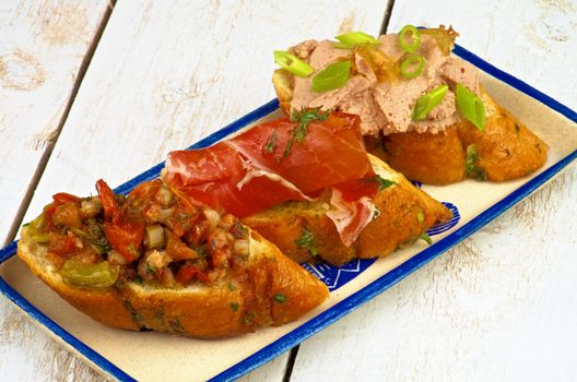 Mix Tapas Bruschetta with Bacon, Roasted Vegetables, Cured Ham and Pate with Spring Onion on Garlic Bread closeup on Plate on White Wooden background