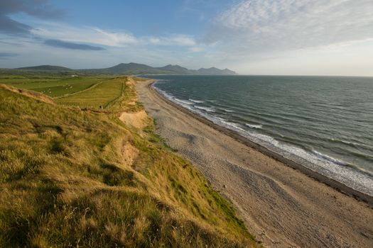 Grasses blowing in the wind lead to green farm land and sand banks in the evening sun with a pebble and sand beach leading to mountains in the distance. Dinas Dinlle, Wales coast path, Gwynedd, Wales, UK