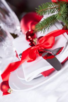 Romantic red Christmas table setting with white plates, red and white linen and silverware tied with a red ribbon and bow decorated with red Xmas baubles and evergreen natural pine foliage