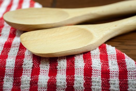 Two wooden spoons sit on a red and white cloth and a wooden table.