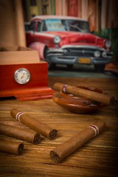 Cuban cigars and humidor with ashtray on rustic wooden table with Cuban painting of american old car in background