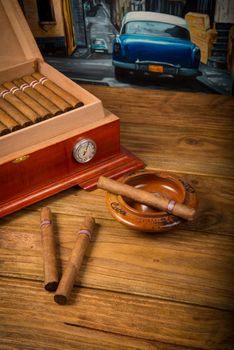 Cuban cigars and humidor with ashtray on rustic wooden table with Cuban painting of american old car in background