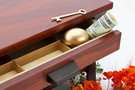 Feminine, financial image of savings depicted with wood grain box, open drawer, gold nest egg, key, and American currency with flowers visible below. 