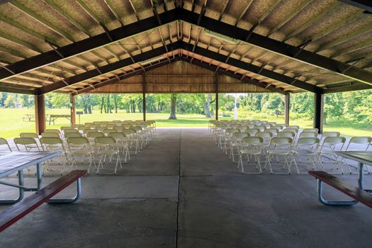 A picnic shelter set up for a wedding ceremony with many rows of white folding chairs. Shot from behind. 