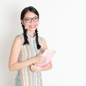 Portrait of Asian Chinese school girl hands holding text books in retro revival style cheongsam standing on plain background.