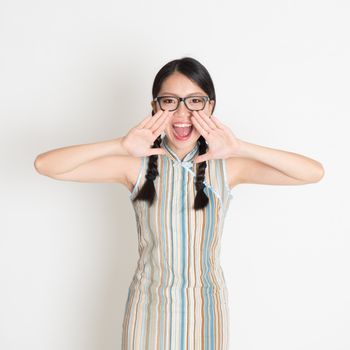 Portrait of Asian Chinese girl  shouting loud, hands next to the mouth, in retro revival style cheongsam standing on plain background.