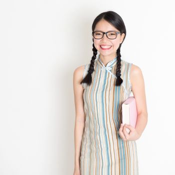 Portrait of Asian Chinese college girl hands holding text books in retro revival style cheongsam standing on plain background.