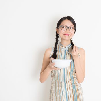 Portrait of Asian Chinese woman eating, using chopsticks holding rice bowl, in retro revival style cheongsam standing on plain background.