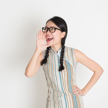 Portrait of Asian Chinese female  shouting loud, hand next to the mouth, in retro revival style cheongsam standing on plain background.