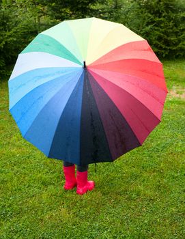 Little girl wearing red rubber boots hiding behind colorful umbrella in the rain