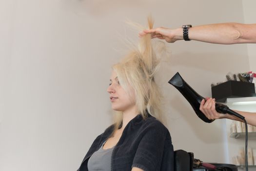 Beautiful blonde girl sitting, getting hair dried. Hands of hairdresser visible, holding blow dryer