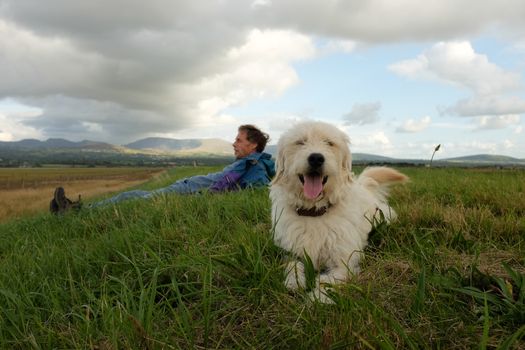 A labradoodle, dog, lays on green grass looking forward with the owner, a man, lays on the grass behind.