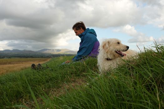 A labradoodle, dog, lays on green grass looking to one side, with the owner, a man, sitting on the grass behind.