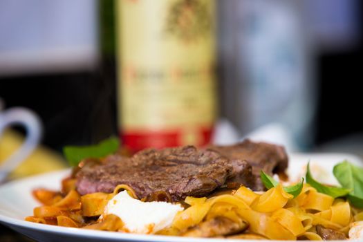 Serving of beef steak with Italian pasta on a plate in a restaurant or on a dinner table, low angle view with a bottle of wine behind