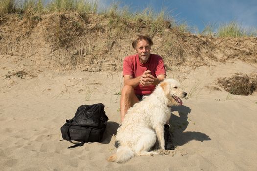 A man and dog, labradoodle, sitting on a beach under a sand dune staring ahead.