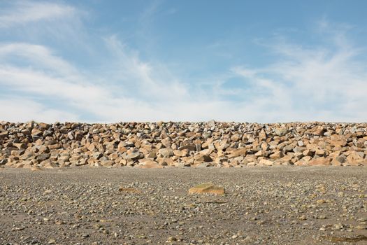 Sand and pebbles lead to a wall of rocks and boulders, sea defences, with a blue sky with cloud in the distance.