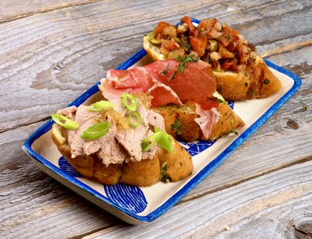 Arrangement of Tapas Bruschetta with Pate and Spring Onion, Bacon, Roasted Vegetables and Cured Ham on Garlic Bread on Plate on White Wooden background