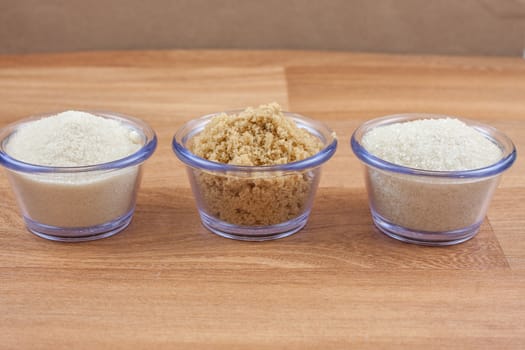 Organic crystal, brown, and fine sugar in clear bowls on a wooden surface.