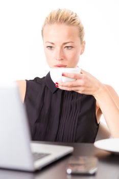 Business woman holding white cup of coffee in her hand while checking some information on her laptop computer in office. Multitasking.