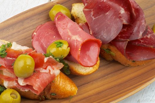 Delicious Tapas with Smoked Jamon, Cured Beef, Garlic Bread and Green Olives closeup on Wooden Plate 