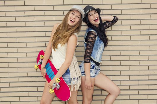Two beautiful and young girlfriends having fun with a skateboard, in front of a brick wall