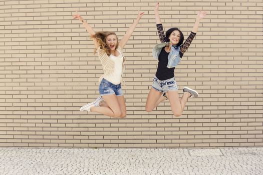 Two beautiful and young jumping in front of a brick wall