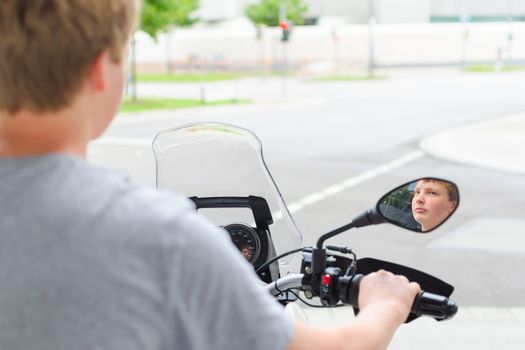 Teenager riding a motorcycle and stopped at a red light of the street junction. Small DOF with face reflection in the mirror.