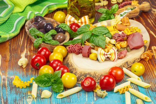 Fresh ingredients for making pasta dish with a herbs, olive oil, olives, tomatoes and uncooked pasta different colors.