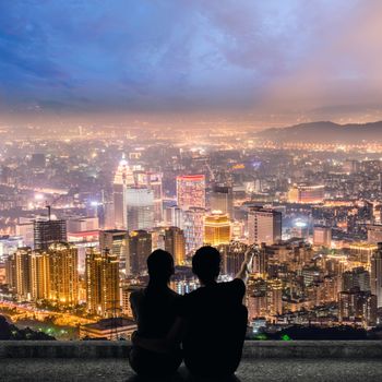 Silhouette of couple sit on ground point faraway on the roof above the city in the night.