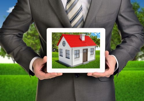 Hands hold tablet pc. Picture of small house with red roof on screen. Blurred landscape as backdrop