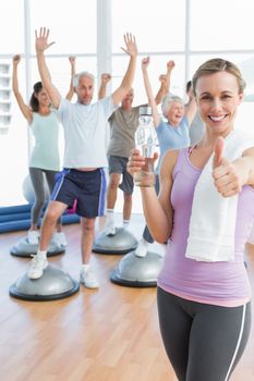 Young woman gesturing thumbs up with people stretching hands in the background at fitness studio