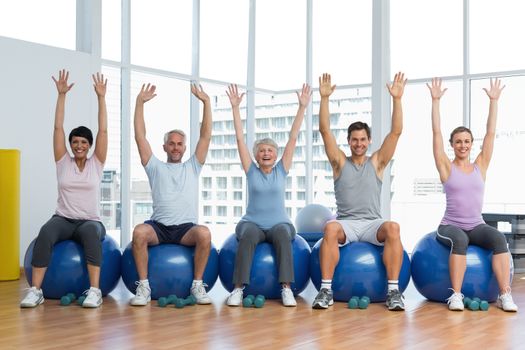 Portrait of fitness class sitting on exercise balls and raising hands in a bright gym