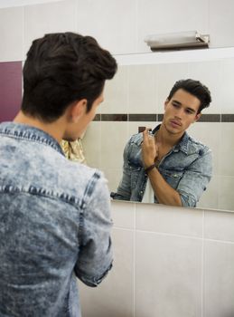 Attractive young man looking at bathroom mirror, spraying cologne or perfume