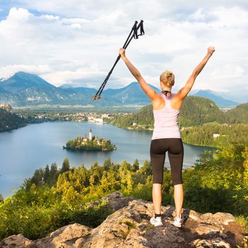 Made it! Young spoty active lady with hiking sticks and hands rised admiring beautiful nature around Bled Lake in Julian Alps, Slovenia. 