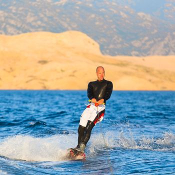 Wakeboarder in wetsuit riding in sunset. Wakeboarding is a surface water sport which involves riding a wakeboard over the surface of a body of water.