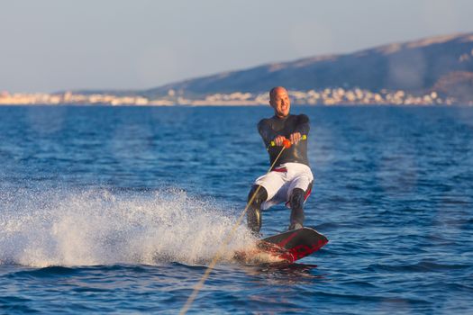 Wakeboarder in wetsuit riding in sunset. Wakeboarding is a surface water sport which involves riding a wakeboard over the surface of a body of water.