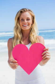 Smiling blonde showing pink heart on the beach on a sunny day