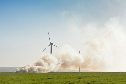 A small wind farm in a green field with a controlled fire burning close by. Thick clouds of smoke conceal most of the nearest turbines.