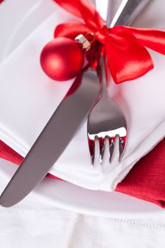 Romantic red Christmas table setting with white plates, red and white linen and silverware tied with a red ribbon and bow decorated with red Xmas baubles and evergreen natural pine foliage