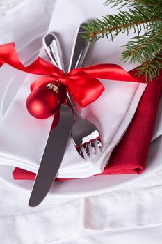 Romantic red Christmas table setting with white plates, red and white linen and silverware tied with a red ribbon and bow decorated with red Xmas baubles and evergreen natural pine foliage