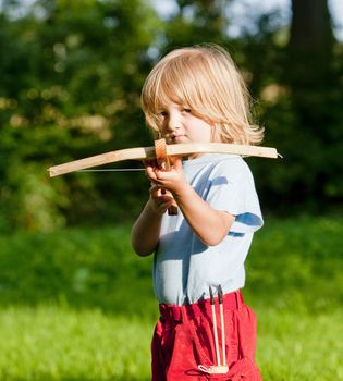 little boy with long blond hair shooting with toy crossbow in the garden