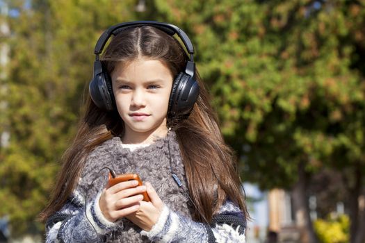 Beautiful little girl listening to music on headphones in autumn park