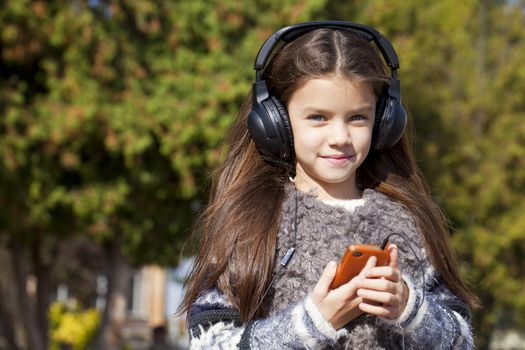 Beautiful little girl listening to music on headphones in autumn park