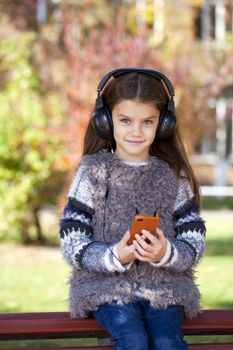 Beautiful little girl listening to music on headphones in autumn park