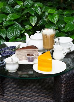 Coffee, tea, chocolate crape cake and orange cake on table in outdoor garden