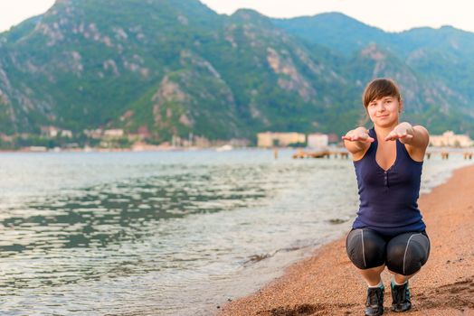 beautiful girl performs squats on the sand near the sea