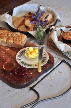 Freshly baked products laid out for breakfast with butter buttons and jam. Wholegrain bread on a cutting board, muffins and white bread in baskets.