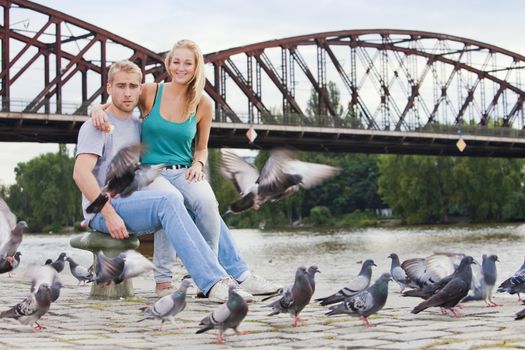 young happy couple sitting by the riverside, feeding birds