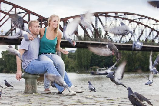 young happy couple sitting by the riverside, feeding birds